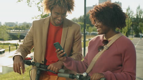 Amigos-Afroamericanos-Positivos-Parados-Al-Aire-Libre-Con-E-scooters-Y-Usando-El-Teléfono
